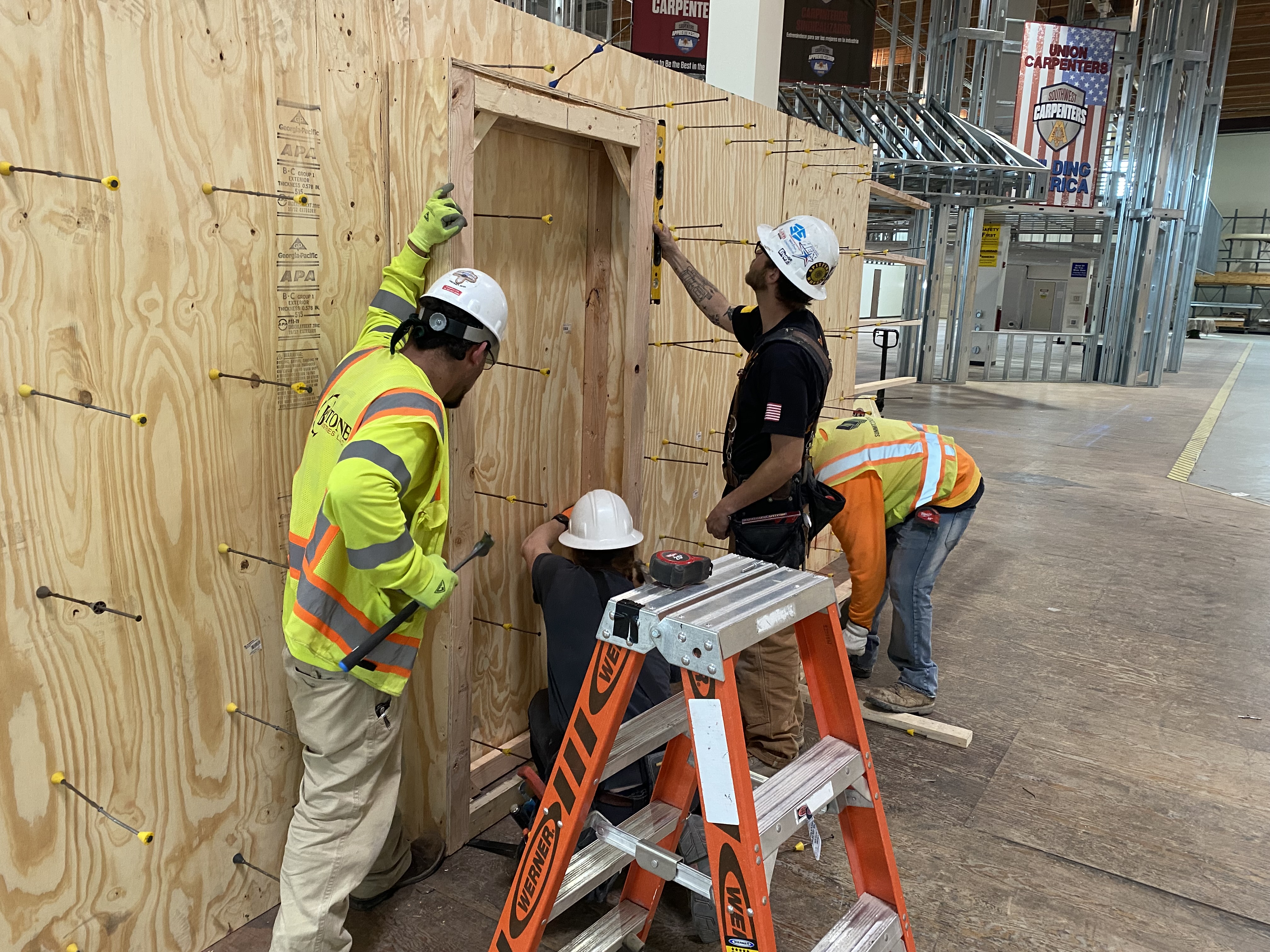 Four carpenters install a doorframe on a plywood structure. 